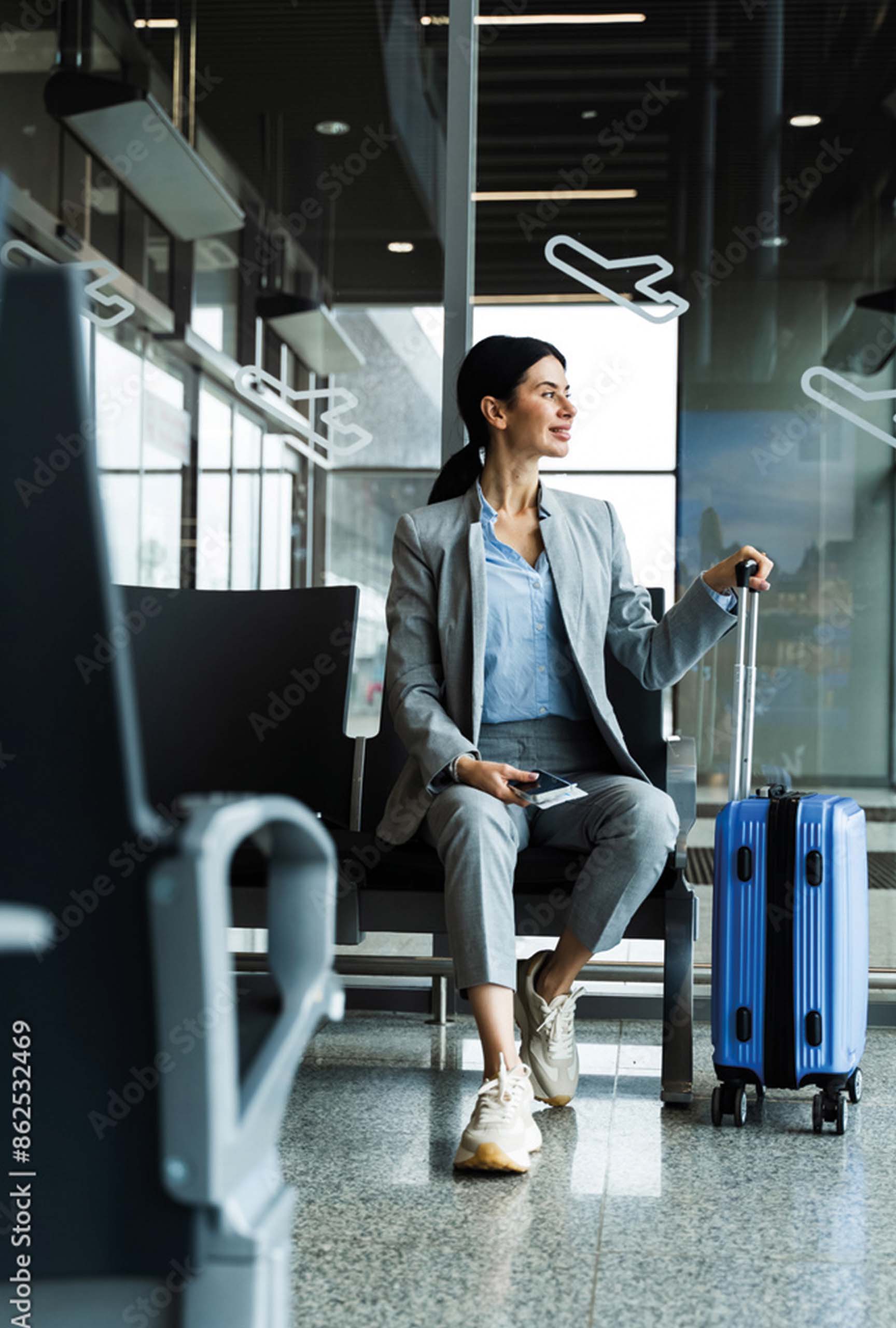 Person im Flughafen‑Terminal, sitzend mit Rollkoffer, Smartphone und Reisedokumenten vor großen Glasflächen mit Flugzeugsymbolen.