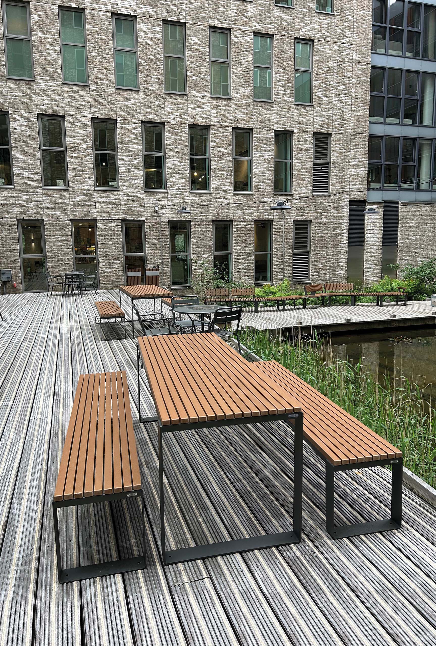 Moderner Außenbereich mit Holzterrasse, Outdoor‑Sitzgruppen und einem kleinen Wasserbecken vor einem Backsteingebäude.