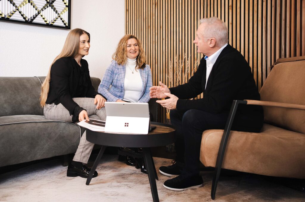 Drei Personen sitzen in einem modernen Besprechungsraum zu einem Gespräch zusammen – zwei Frauen auf einem Sofa, ein Mann im Sessel gegenüber.