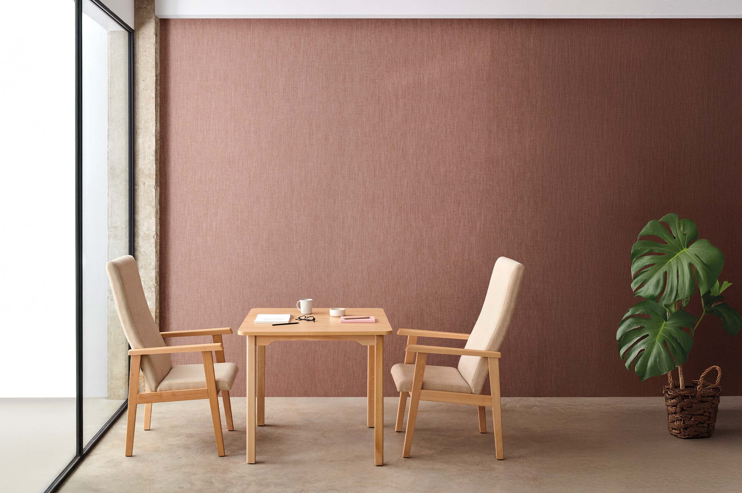 Minimalistisch eingerichteter Innenraum mit kleinem Holztisch, zwei gepolsterten Stühlen, Notizbüchern und einer großen Monstera‑Pflanze.
