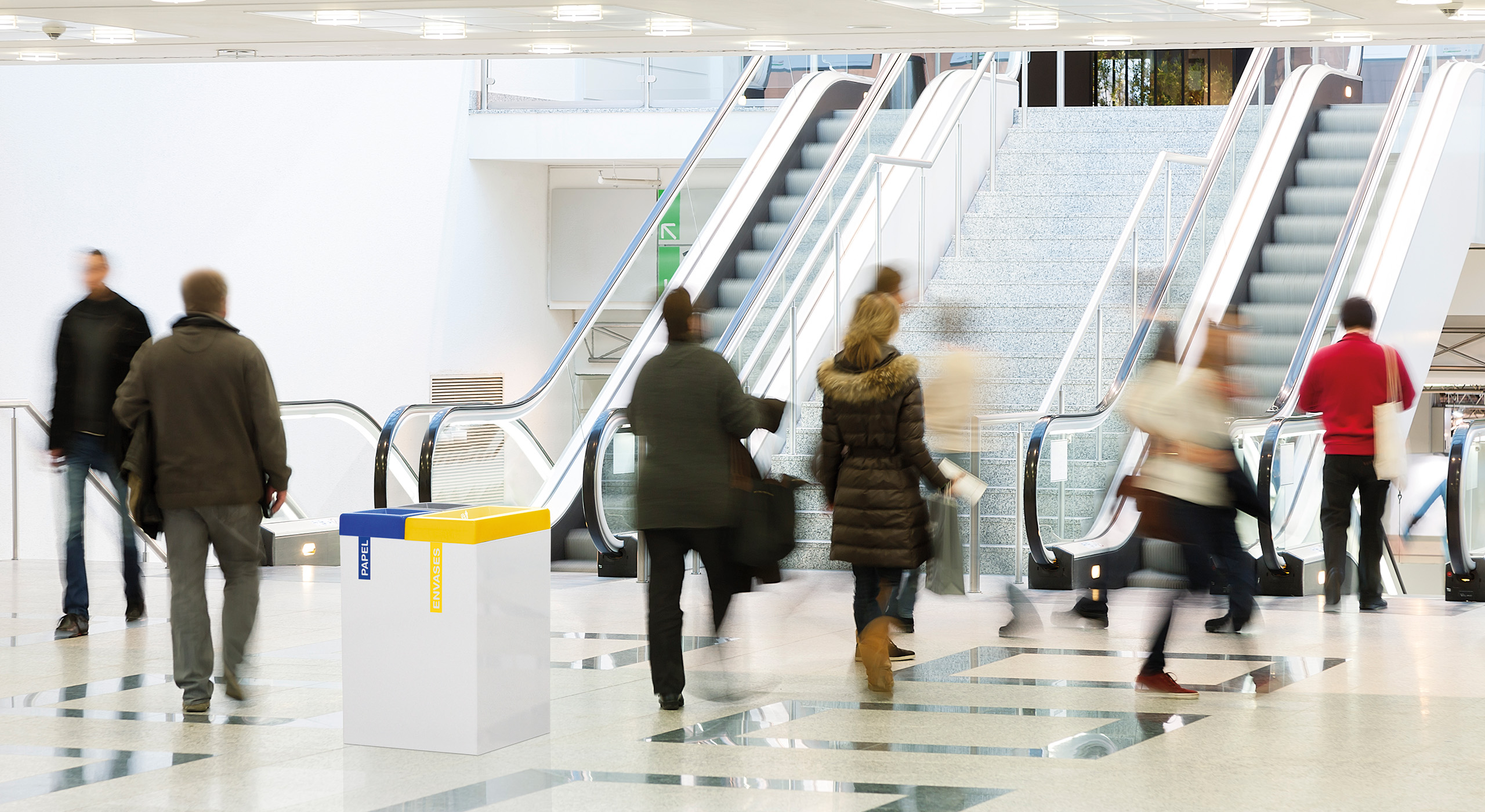 Recyclingbehälter in einem hellen, modernen Innenraum mit Rolltreppen und vorbeigehenden Menschen.